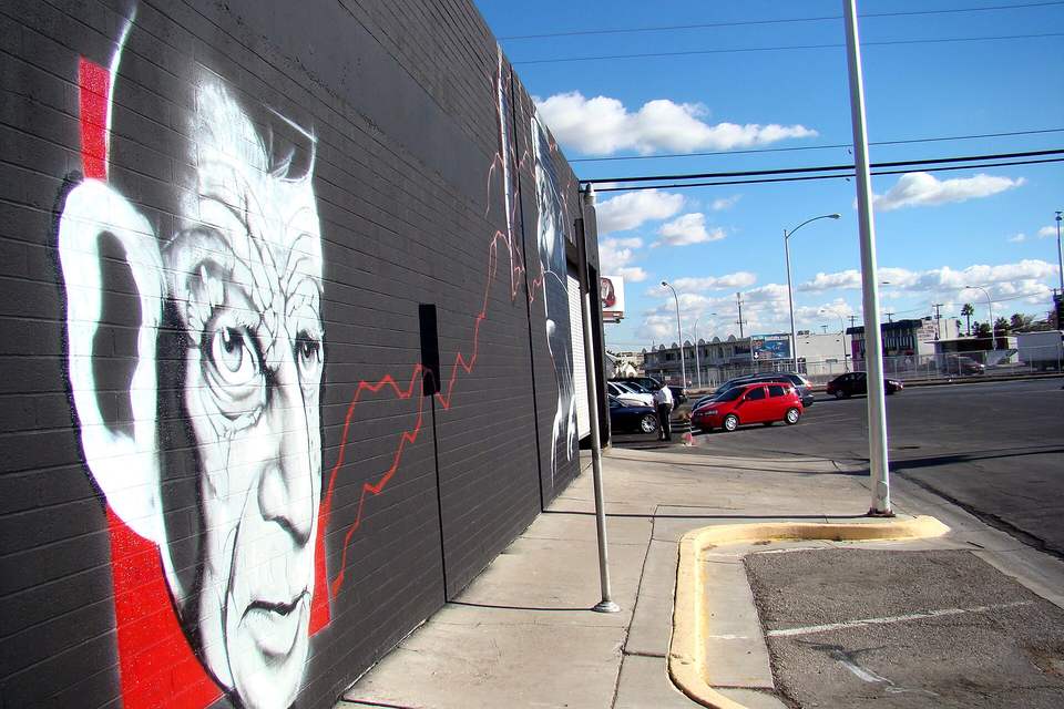 Large black and white portrait mural on a brick building beside a sidewalk and parking lot under a blue sky