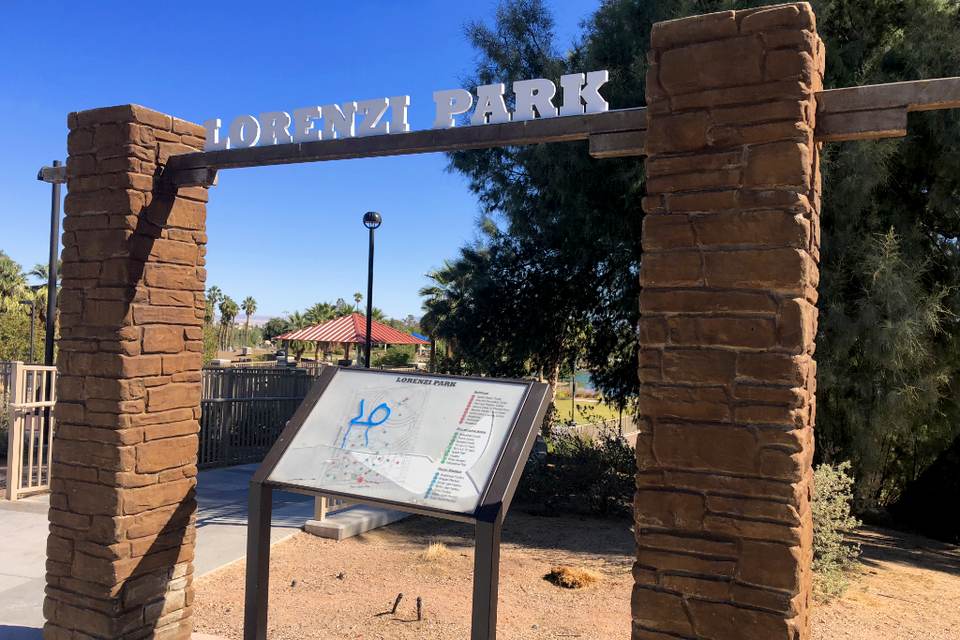 Stone pillar gateway with a Lorenzi Park sign, a map board and trees behind it